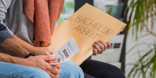 Bachelorarbeit Zwei Personen sitzen nebeneinander. Man sieht einen Ausschnitt von Knie bis Brust. Die hintere Person hat einen großen Umschlag mit der Aufschrift „Bachelor-Arbeit“ in der Hand, die vordere Person hat einen kleinen weißen Zettel mit einer Wartenummer, einem Code und dem TU-Logo in der Hand.